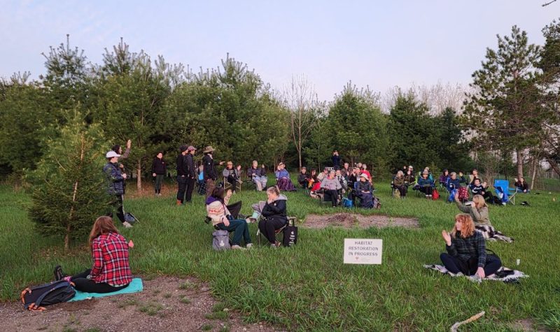 Early morning. A group of over 50 people sit on blankets and lawn chairs in a grassy area surrounded by trees.