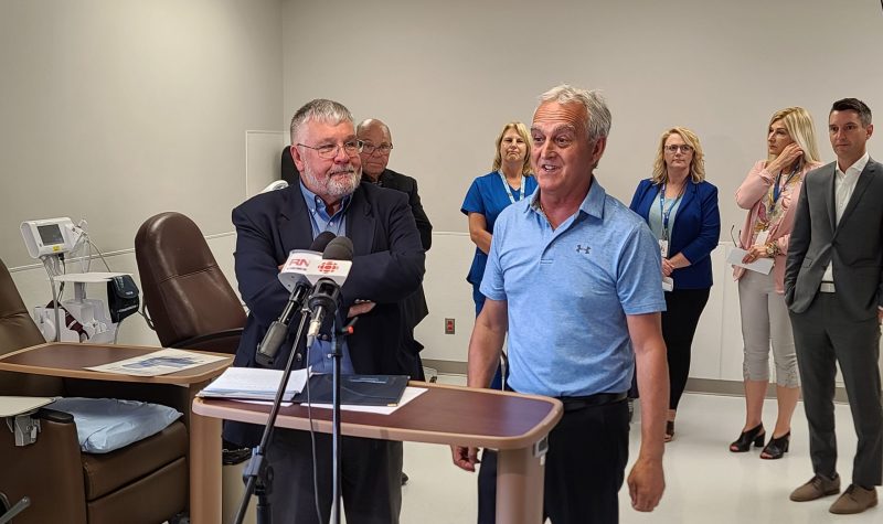 Two men stand in front of a podium in an oncology unit at a hospital, with several other people standing in the background.