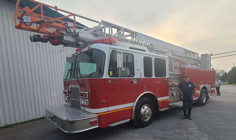 A man in uniform poses with a new fire truck with a large ladder in a bare parking lot.