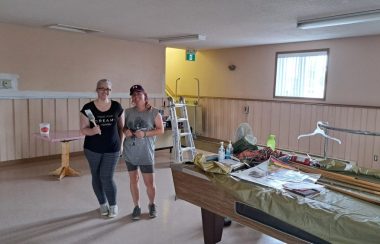 Deux femmes à l'intérieur d'une salle tiennent un pinceau à la main et s'apprêtent à peinturer la salle devant un table de billard contenant plusieurs outils de peinture.