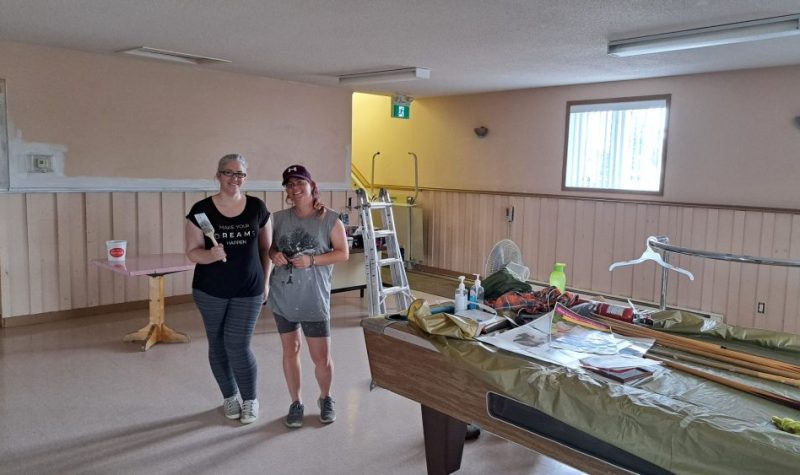 Deux femmes à l'intérieur d'une salle tiennent un pinceau à la main et s'apprêtent à peinturer la salle devant un table de billard contenant plusieurs outils de peinture.