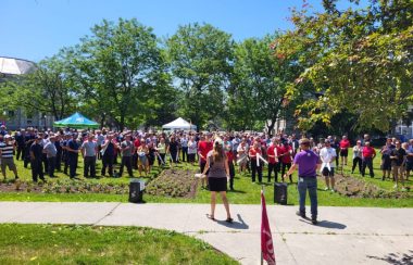 A large group of people gather in a field on Queen's campus. There are many large green trees surrounding the area behind the crowd. There are a few tents for shade and it is a sunny day with blue skies.
