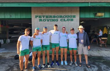 Head coach and 6 rowers stand in front of Peterborough Rowing Club.