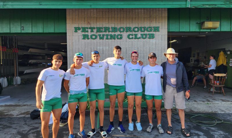 Head coach and 6 rowers stand in front of Peterborough Rowing Club.
