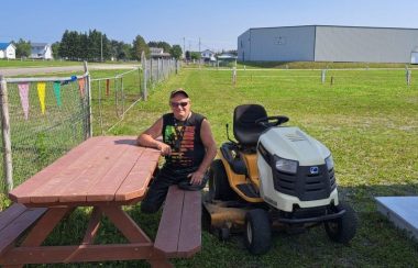 Un homme amputé des deux jambes est assis sur une table de pique nique, au côté de son tracteur à pelouse, sur un vaste terrain de pelouse, lors d'une journée chaude et ensoleillée