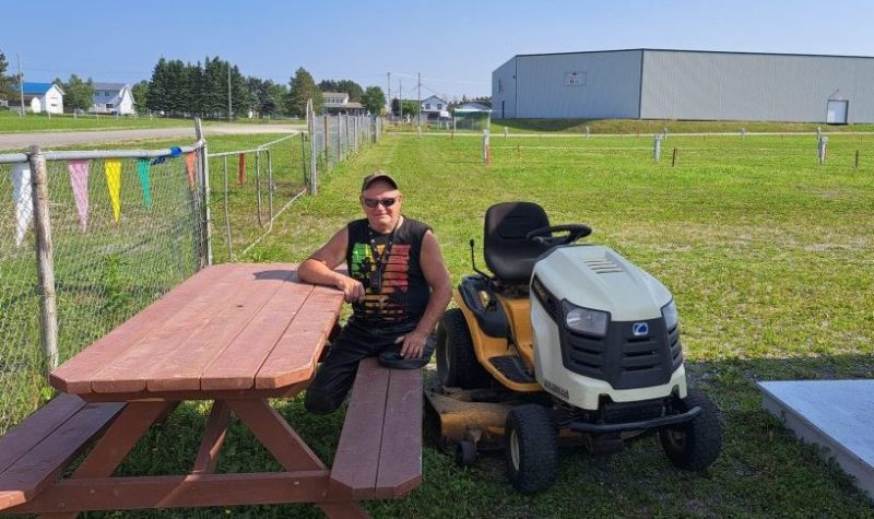 Un homme amputé des deux jambes est assis sur une table de pique nique, au côté de son tracteur à pelouse, sur un vaste terrain de pelouse, lors d'une journée chaude et ensoleillée