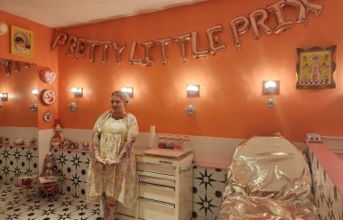 Jessie Darling standing in a piercing studio, decorated in bright pinks and oranges, with metallic surfaces and loud designs.