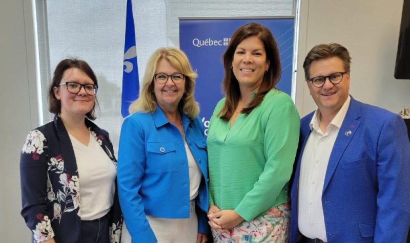 Trois femmes et un homme en rang prenant la pose devant un drapeau du Québec