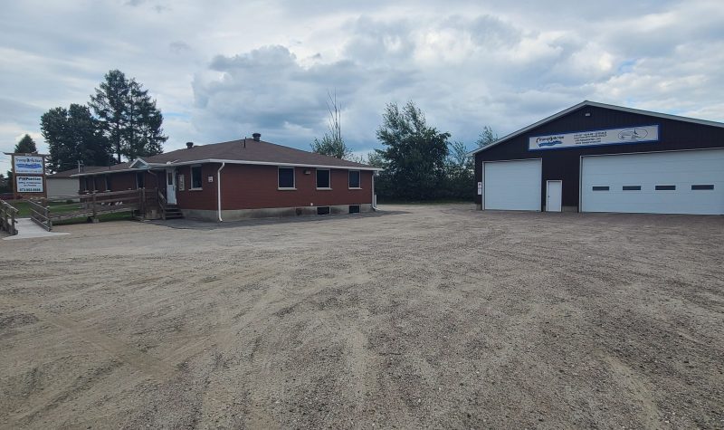 A red brick low rise building and a grey and white two door garage sits on a large gravel driveway.