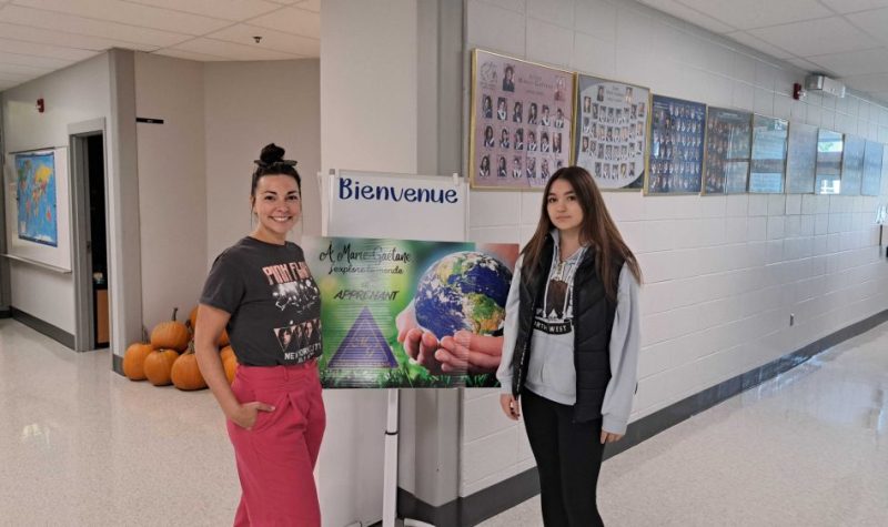 de gauche à droite, une enseignante et une élève devant l'affiche dévoilant la thématique de l'année scolaire 2023 : À Marie-Gaétane, j'explore le monde en apprenant.