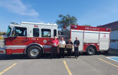 Fire truck behind 2 members of council, the fire chief, and a representative from Enbridge Gas holding boxes of fire alarms.