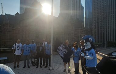 A group of Toronto Argonauts players and mascot with Mayor Oliva Chow raise a flag