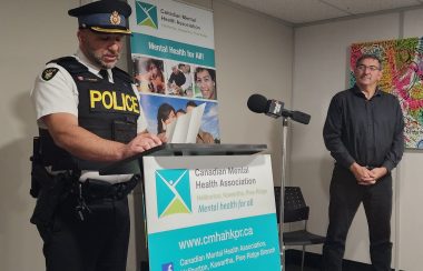 A man in a police uniform another in black shirt and pants stand next to a turquoise and white banner containing the words 'Canadian Mental Health Association'.