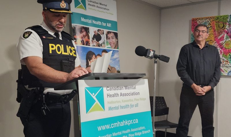 A man in a police uniform another in black shirt and pants stand next to a turquoise and white banner containing the words 'Canadian Mental Health Association'.