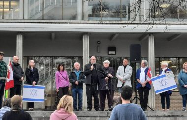 A crowd is assembled at the top of Prince George City Hall's step. A man speaks into the microphone flanked by the city's mayor. Flags of Israel and Canada can be seen.