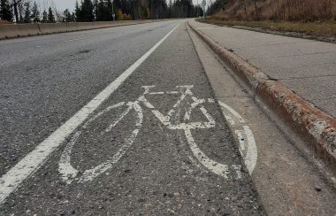 We look up a gradual hill ata paved road surface. PaintedA narrow shoulder of a paved road with the image of a bicycle. The front wheel touches the painted road margin, and the back wheel is in the rain gutter.