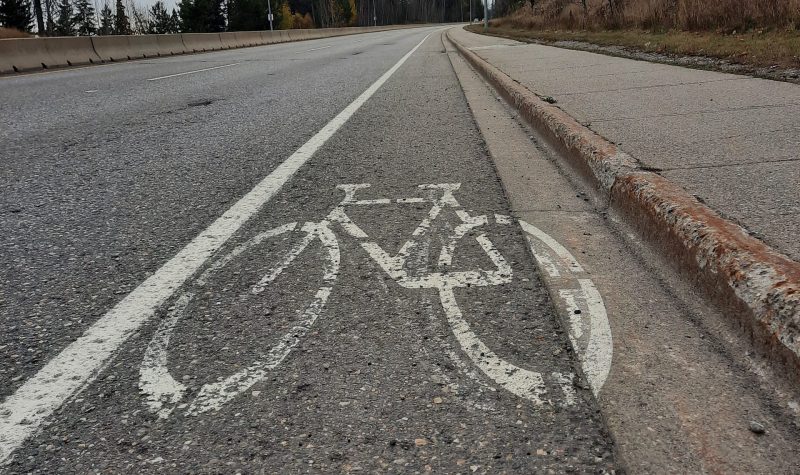 We look up a gradual hill ata paved road surface. PaintedA narrow shoulder of a paved road with the image of a bicycle. The front wheel touches the painted road margin, and the back wheel is in the rain gutter.