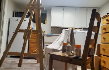 An small art studio with an easel and two chairs. One with paint supplies and the other with a cloth over it.