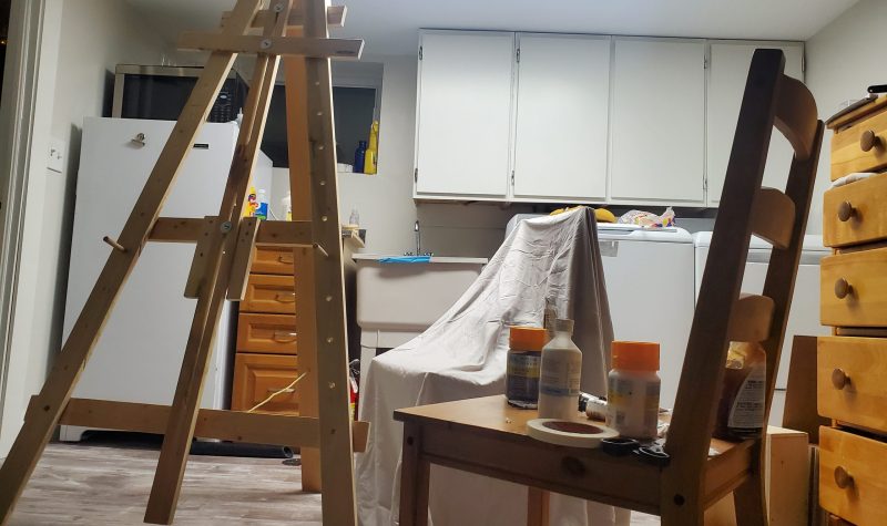 An small art studio with an easel and two chairs. One with paint supplies and the other with a cloth over it.
