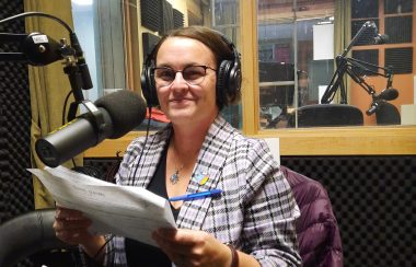 A woman is seated in front of a microphone boom in a recording studio. She wears a purple plaid shirt, headphones, glasses, and brown hair. Her purple jacket is draped over her chair.