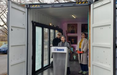 Councillor Alejandra Bravo behind a podium in a shipping container. With two other people behind her and art on the wall.