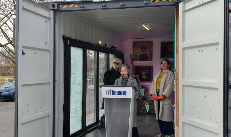 Councillor Alejandra Bravo behind a podium in a shipping container. With two other people behind her and art on the wall.