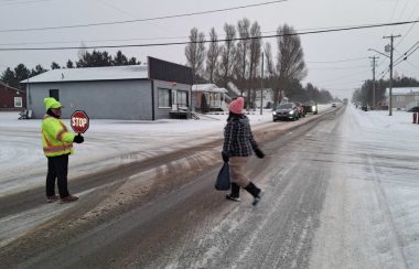 une brigadière tenant une pancarte d'arrête aidant une écolière à traverser une rue enneigée. Des véhicules sont arrêtés pour laisser passer l'écolière, lors d'une journée de novembre nuageuse et enneigée