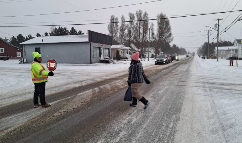 une brigadière tenant une pancarte d'arrête aidant une écolière à traverser une rue enneigée. Des véhicules sont arrêtés pour laisser passer l'écolière, lors d'une journée de novembre nuageuse et enneigée