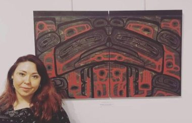 A woman stands next to a red and black Indigenous art work on a wall