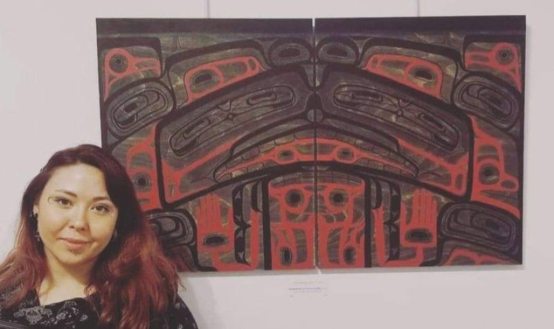 A woman stands next to a red and black Indigenous art work on a wall