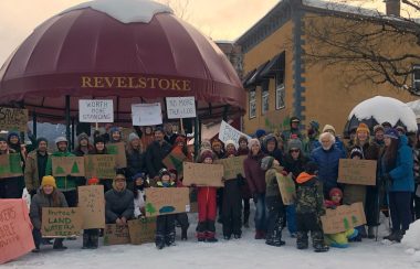 A crowd groups together for a photo holding protest signs under a gazebo that says Revelstoke on it.