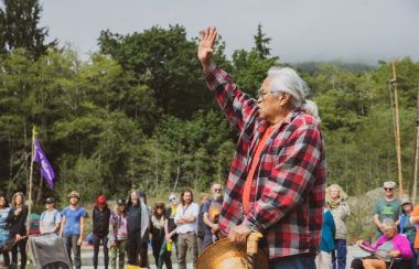 A First Nations raises his hand while addressing a group of young people in the woods