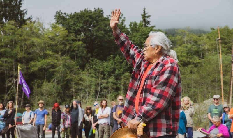 A First Nations raises his hand while addressing a group of young people in the woods
