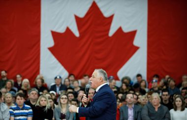 Darrell Samson, un homme en costume grisonnant au micro devant une foule et un grand drapeau canadien.