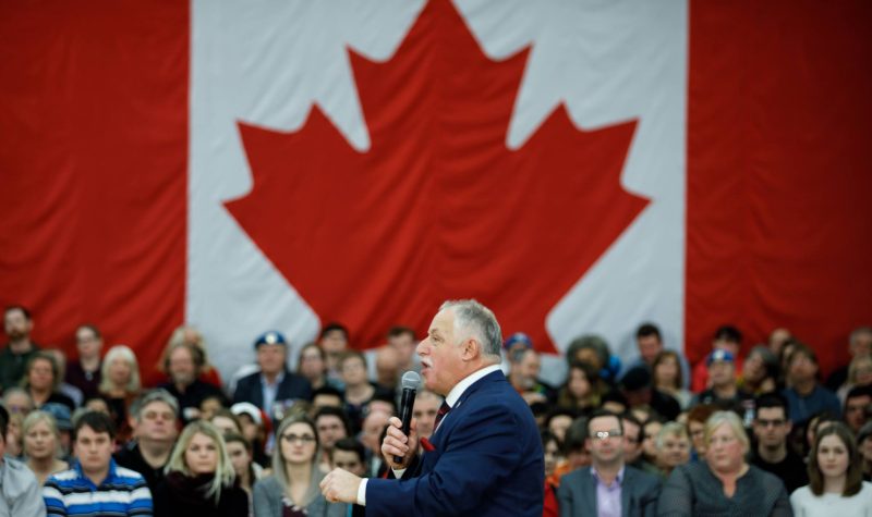 Darrell Samson, un homme en costume grisonnant au micro devant une foule et un grand drapeau canadien.