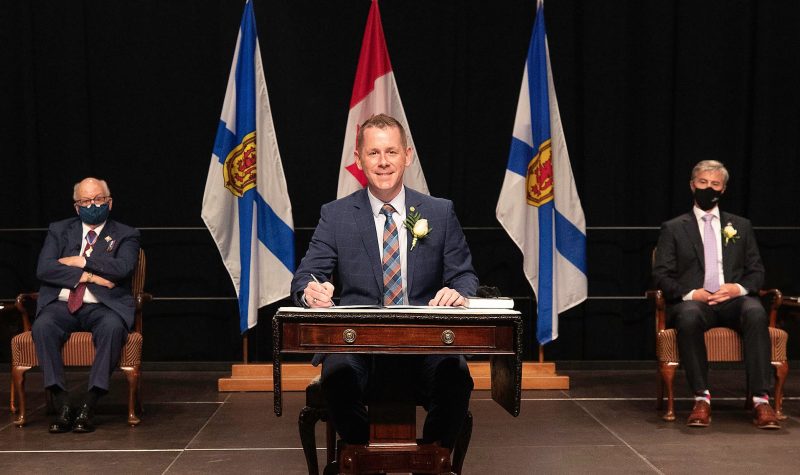 Colton Leblanc au bureau d'investiture, un stylo à la main et souriant pour la photo. A sa droite assis, Tim Houston. Au fond, les drapeaux du Canada et de la Nouvelle-Ecosse.