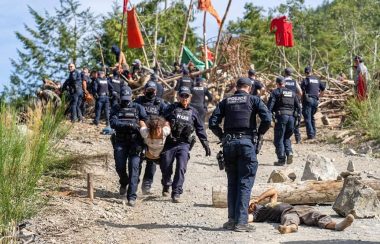 Three police offciers remove a young woman from a blockade. Another officer stands over a male forest defender who is stretched out on the ground. IN the background; police and protesters at the blockade