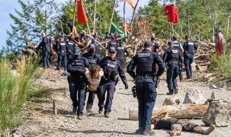 Three police offciers remove a young woman from a blockade. Another officer stands over a male forest defender who is stretched out on the ground. IN the background; police and protesters at the blockade