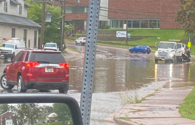 A red SUV is seen on a street that has flooded. There is a truck and a red brick building in the distance.