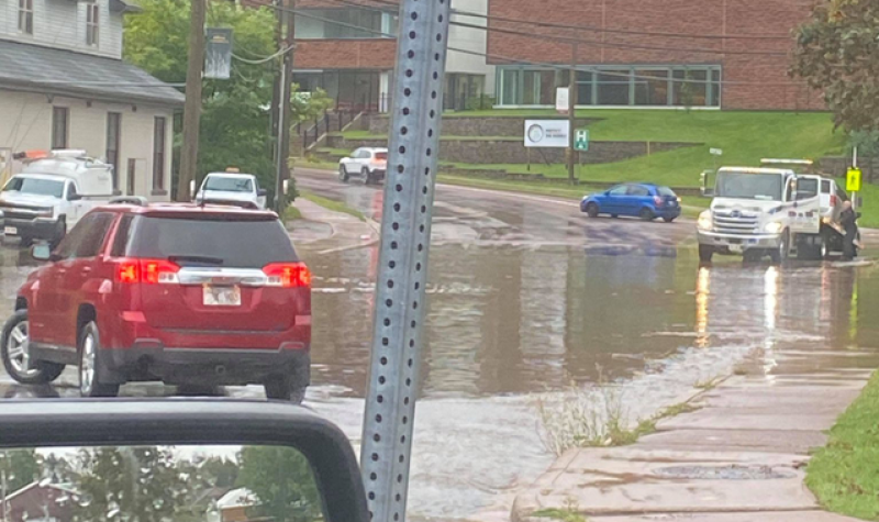 A red SUV is seen on a street that has flooded. There is a truck and a red brick building in the distance.