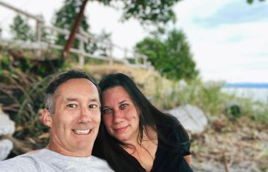 A couple sitting together on the beach. A log, fence and grass covered trail are behind them.