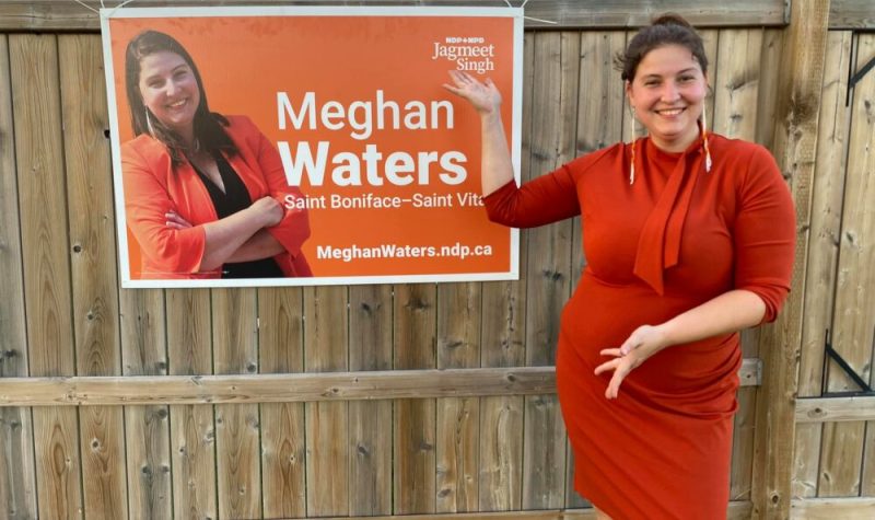 Meghan, souriante et habillée en robe orange, pointe à une pancarte électorale d'elle même accrochée sur une haute clôture en bois.