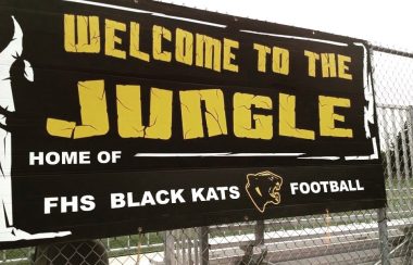 Pancarte accroché sur la grille qui entoure le terrain de football de Fredericton High School. On peut y lire Welcome to the Jungle, Home of the FHS Black Kats Football. en jaune sur fond noir.