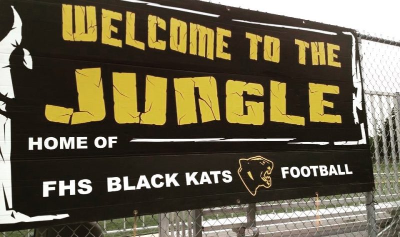Pancarte accroché sur la grille qui entoure le terrain de football de Fredericton High School. On peut y lire Welcome to the Jungle, Home of the FHS Black Kats Football. en jaune sur fond noir.