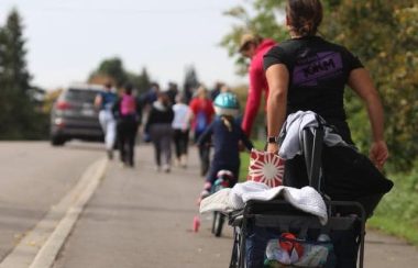 Une femme de dos à la caméra tire une poussette remplie d'objets (couvertures, sacs...). Devant elle, plusieurs personnes courent sur le trottoir, dont un enfant en bicyclette. Il y a une voiture dans la rue et des arbres de l'autre côté du trottoir.