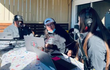 Three indigenous announcers, one female, sitting in front of a building doing live broadcast celebrating the station's first birthday.