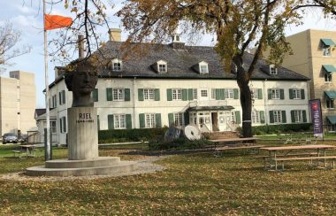 Le musée au mur blanc et fenêtres vertes dans une cours d'herbe pendant l'automne, une statue de Louis Riel devant le musée, et un drapeau orange ballotte au vent à gauche du musée.