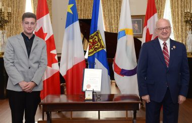 Adrien Comeau a droite, un jeune a lunettes en costume gris pose a coté de la médaille, de l'autre coté, Artur J Leblanc, sourit et pose également, ils sont dans un intérieur richement décoré devant des drapeaux du Canada, de la francophonie, de la nouvelle écosse et de l'Abadie