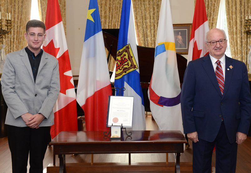 Adrien Comeau a droite, un jeune a lunettes en costume gris pose a coté de la médaille, de l'autre coté, Artur J Leblanc, sourit et pose également, ils sont dans un intérieur richement décoré devant des drapeaux du Canada, de la francophonie, de la nouvelle écosse et de l'Abadie