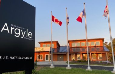 Les bureau municipaux avec devant le panneau de la municipalité et les 4 drapeaux, du Canada, de la Nouvelle-Ecosse, de l'Acadie et du peuple Micmaq. Il fait beau et le soleil est en train de se coucher avec des lumières chaudes